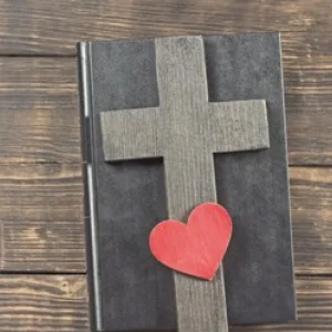 Auf einem, in Leder gebundenem, Buch liegt ein Holzkreuz und ein Herz.